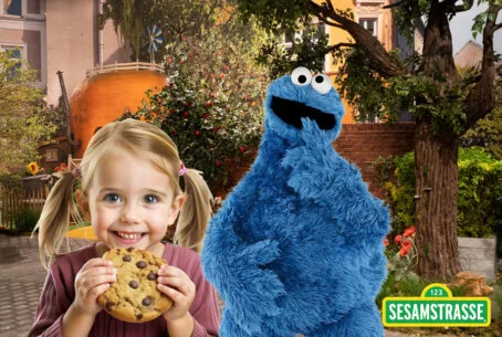 Ein lächelndes kleines Kind mit Zöpfen und einem großen Schokoladenkeks posiert neben der Figur des Krümelmonsters. Das Foto ist mit dem Logo der Sesamstraße versehen.
